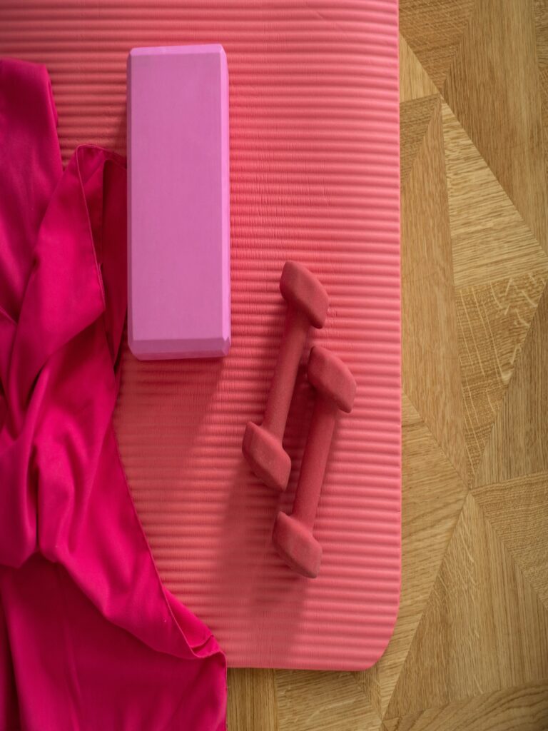 Pink yoga mat, towel, and dumbbells on wooden floor.