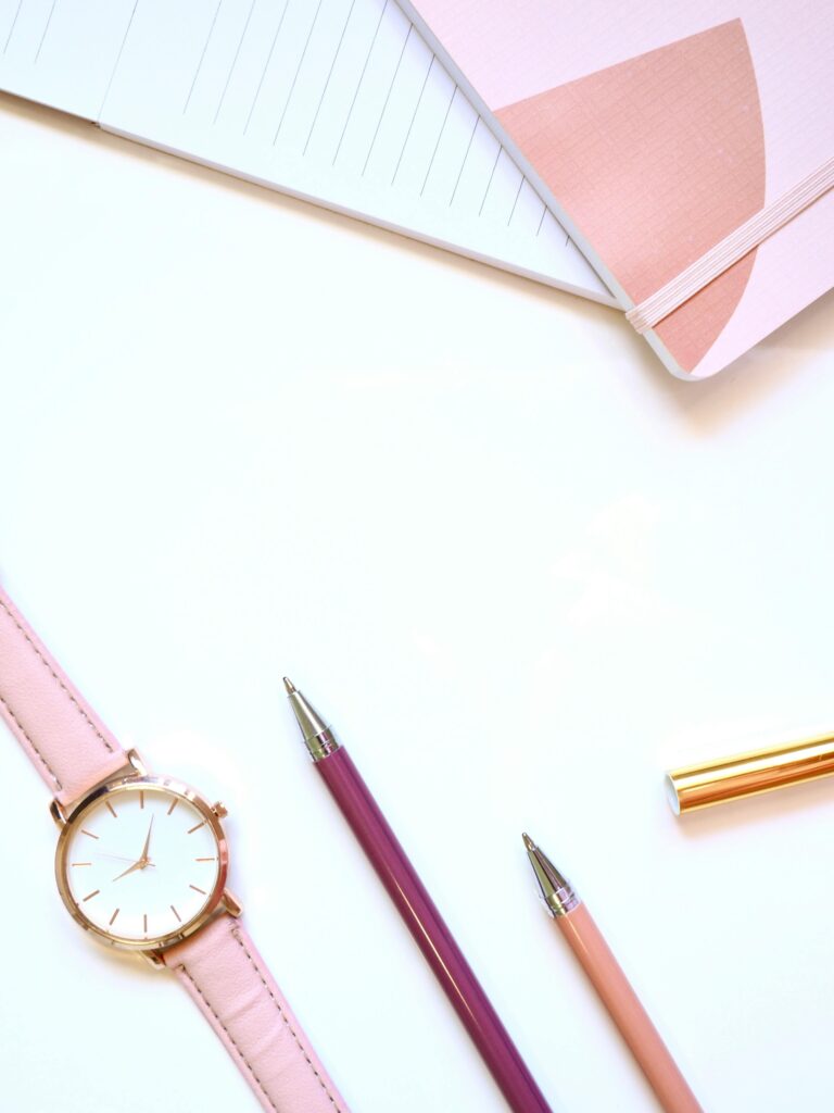 Pink leather wristwatch, pens and notebooks on white surface.