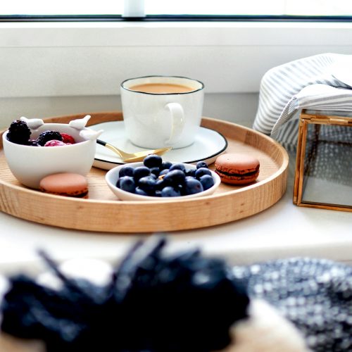 A wooden tray holds a white cup of coffee, a bowl of blueberries, macarons, and berries in a ceramic dish, placed near a window.