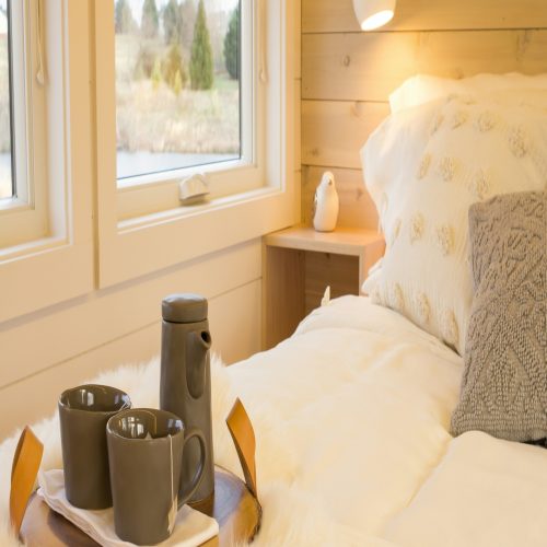 A cozy bedroom with a small wooden tray on the bed holding a dark pitcher and two matching cups.