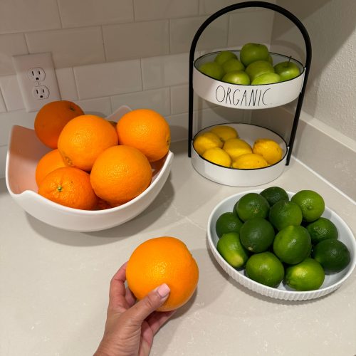 A person holding an orange and a bowl of limes on a wooden table.