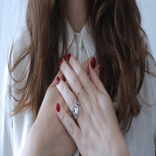 A woman in a white shirt with red nails, holding her hands to her chest.